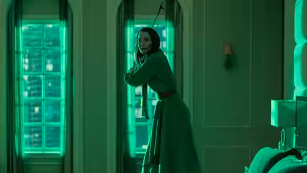 A woman in green coat leans against a doorway in green-lit room