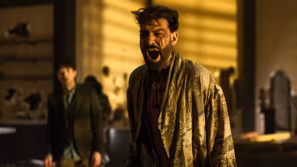 A bloodied man with mouth open stands in a ruined yellow-lit room