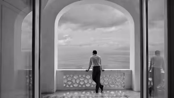 A man stands on a balcony overlooking the sea through an archway