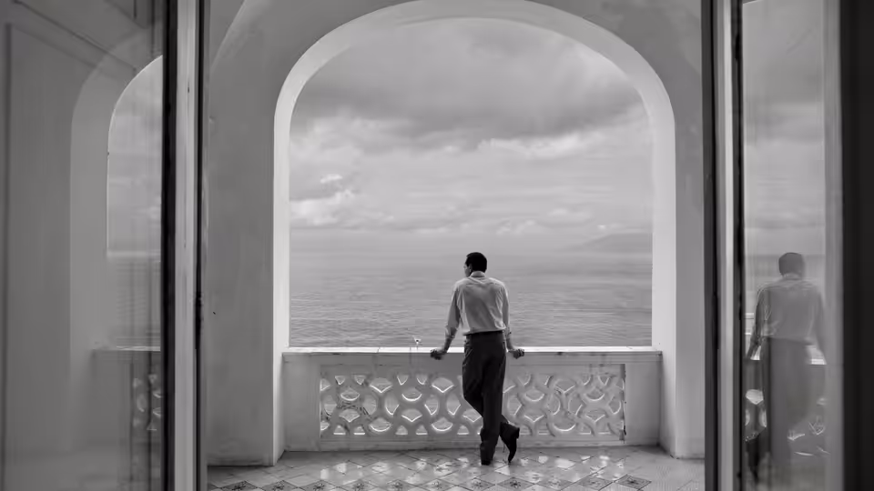 A man stands on a balcony overlooking the sea through an archway