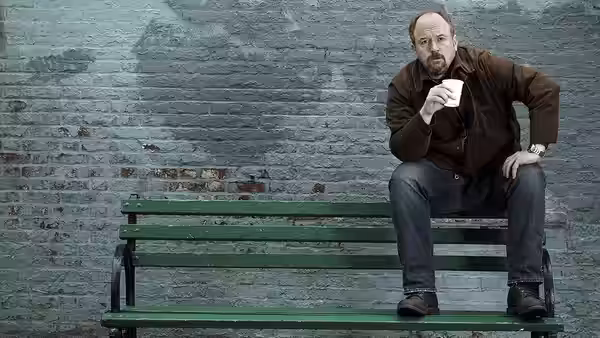 Louie (Louis C.K.) sitting on a green bench holding a paper cup