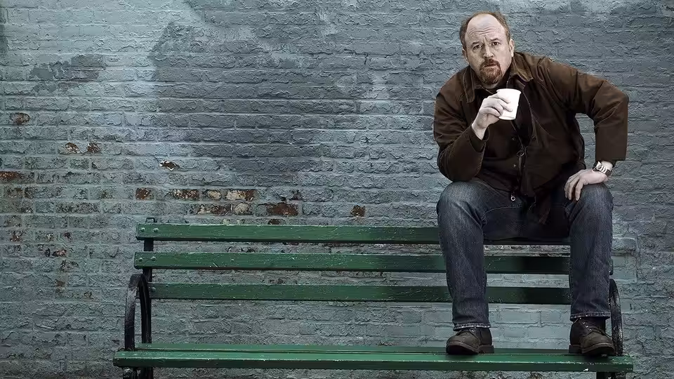Louie (Louis C.K.) sitting on a green bench holding a paper cup