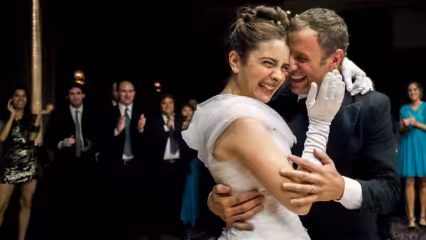 Bride and groom embrace and laugh while guests clap in the background