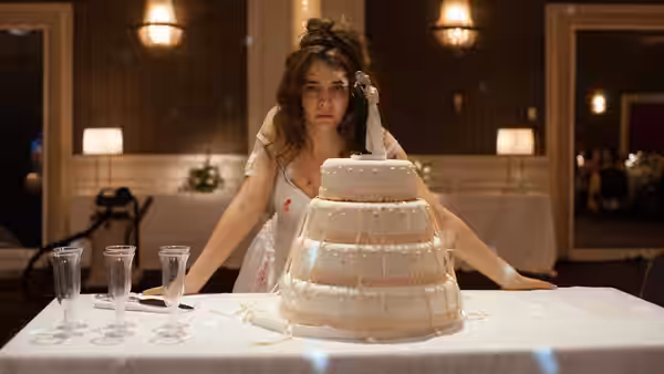 Bride in torn dress leans over a large wedding cake on a table