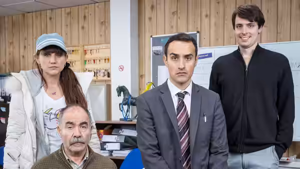 Sophie Charalambos (Natasia Demetriou), Vasos Charalambos (Christos Stergioglou), Stath Charalambos (Jamie Demetriou) and Al stand in an office