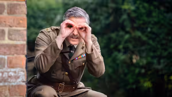 The Captain (Ben Willbond) crouches by a brick wall using his hands as binoculars