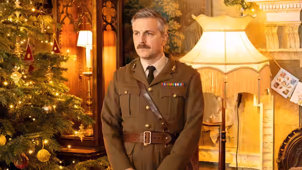 The Captain (Ben Willbond) stands in a living room beside a decorated Christmas tree