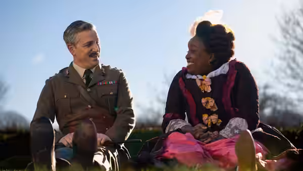 The Captain (Ben Willbond) and Kitty (Lolly Adefope) sit on the grass smiling at each other