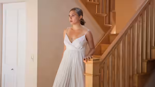 Martha (Elizabeth Olsen) in a white dress stands by a wooden staircase