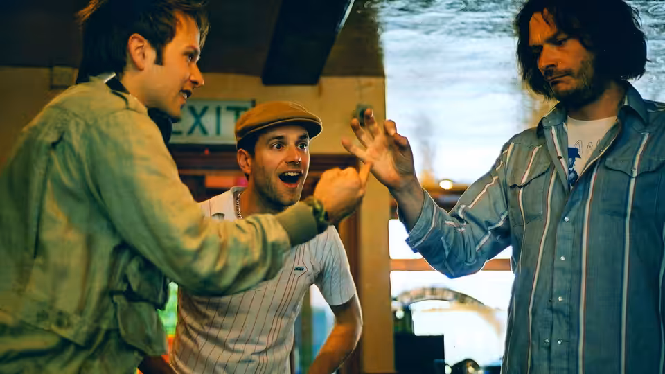Three men gesturing at each other inside a bar near an exit sign