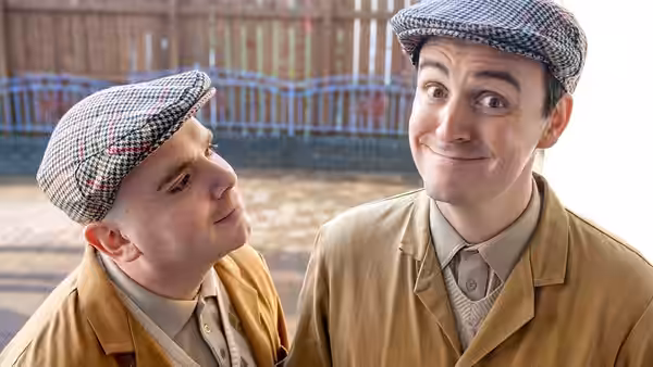 Two men in flat caps and jackets exchange glances outdoors