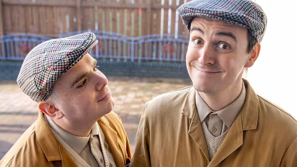 Two men in flat caps and jackets exchange glances outdoors