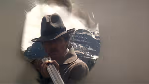 A man in a hat aims a gun through a circular opening at mountains