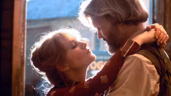 A blonde woman embraces a bearded man with gray hair by a window