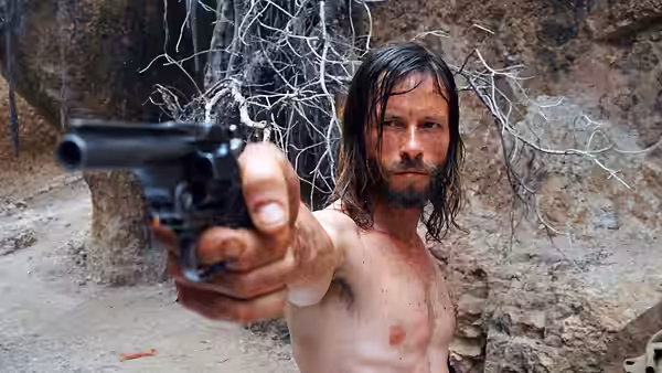 A shirtless, long-haired man aims a pistol in a rocky clearing