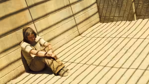 A man sits curled against a wall in a sunlit, barred enclosure
