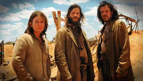 Three men stand among rough wooden structures in a barren camp