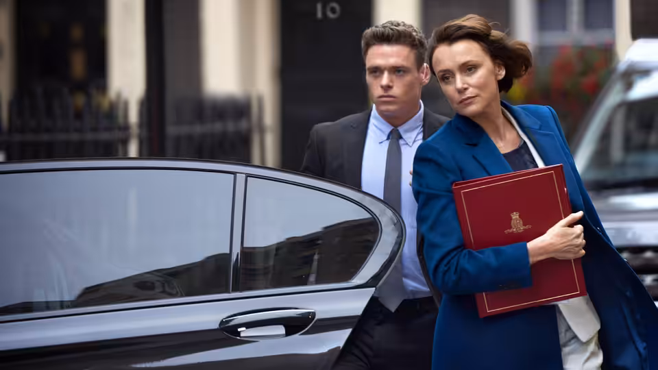 Julia Montague (Keeley Hawes) holding a red folder beside a black car as David Budd (Richard Madden) stands behind her