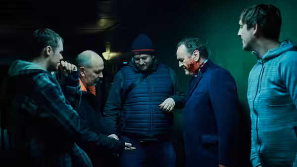 Five men gather closely in a dark room with one face bloodied