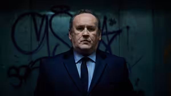 Man in dark suit stands before a wall covered in purple graffiti