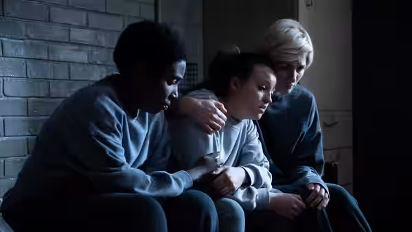 Three women in grey sweatsuits sit closely together against a brick wall