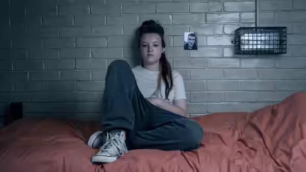 Bella Ramsey as Kelsey Morgan sits cross-legged on a bed in a cell