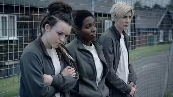 Three women in grey jackets stand by a chain-link fence looking ahead