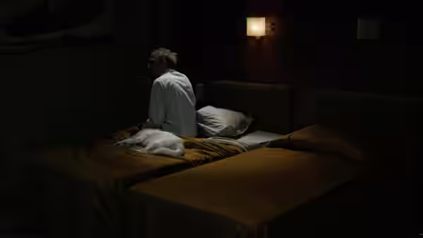 man in white shirt sits on edge of bed in dark room with two beds