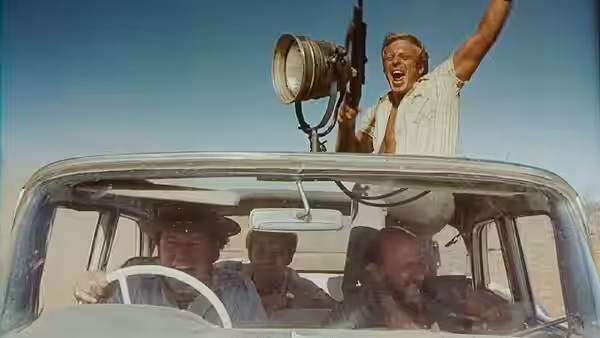 Three men in a car, one standing outside shouting with a rifle raised
