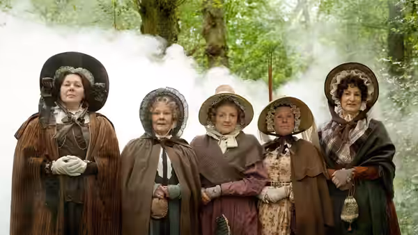 Five women in bonnets and cloaks standing in a line in a misty forest