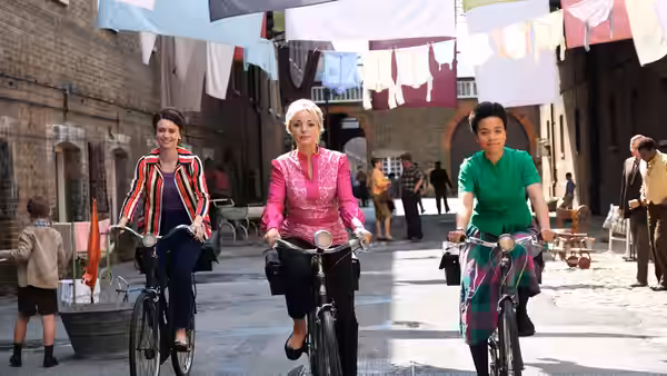 Three women riding bicycles through a street with hanging laundry