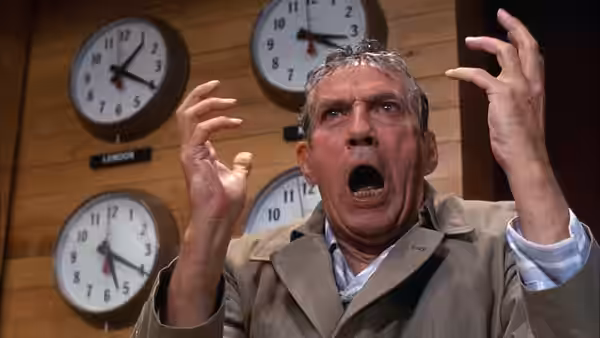 Howard Beale (Peter Finch) with arms raised before several clocks