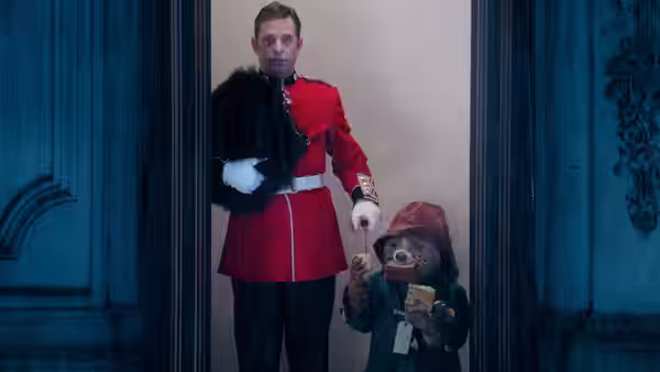 A man in a red uniform stands beside Paddington in a doorway