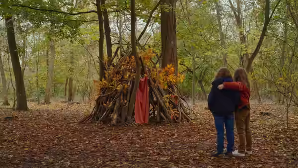 Two girls embrace beside a branch shelter in an autumn forest