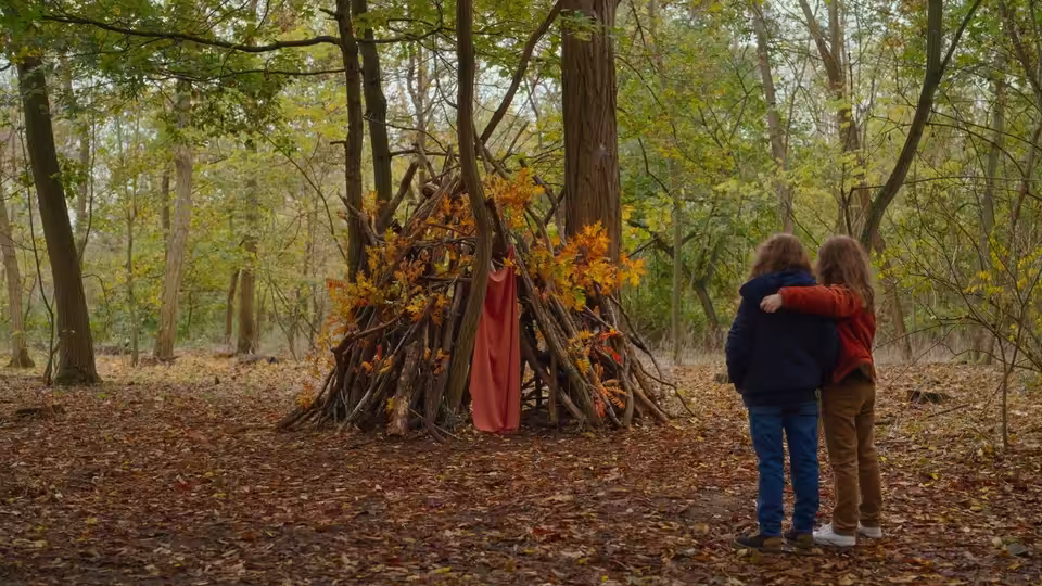 Two girls embrace beside a branch shelter in an autumn forest