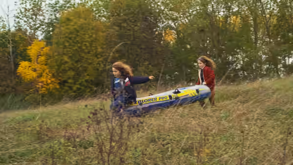 Two girls carry an inflatable raft through a grassy clearing