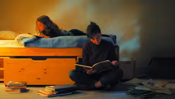A woman reads on the floor beside a girl resting in bed