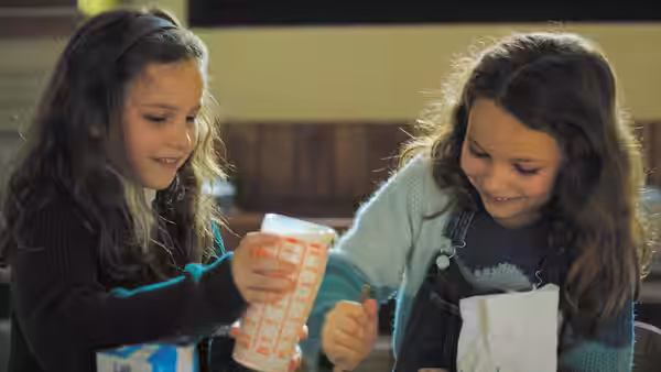 Two girls smile as they prepare food at a kitchen table