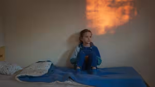 A young girl sits on a bed beneath a patch of warm light