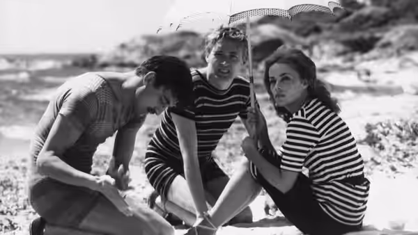 Two men and Catherine gather beneath a beach parasol