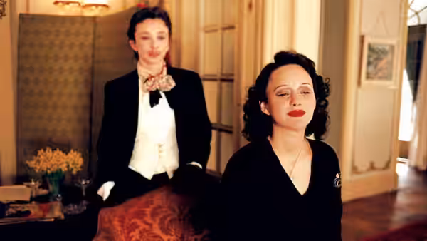 Edith Piaf (Marion Cotillard) sits indoors as another woman stands behind her