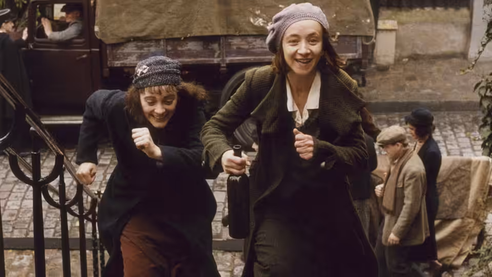 Two young women run up a cobbled street past a horse-drawn carriage