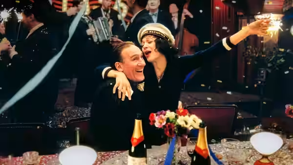 A singer embraces a man at a banquet table amid musicians and champagne
