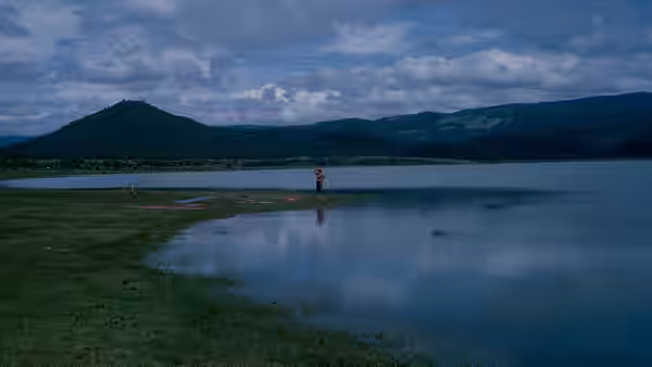 A lone figure stands beside a lake beneath distant mountain ridges