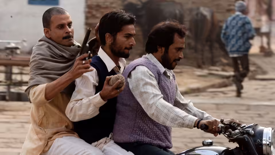 three men riding a motorcycle on a village street one holding a gun