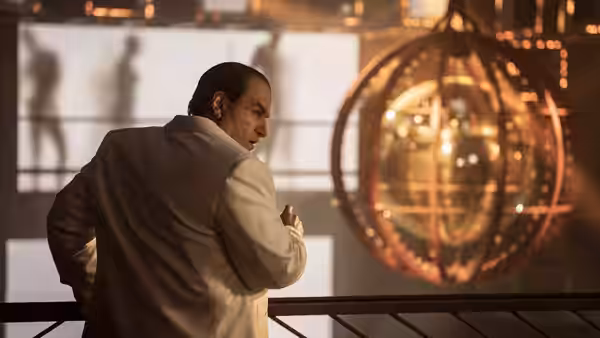 Oz Cobb in a white suit stands before a large glowing spherical chandelier