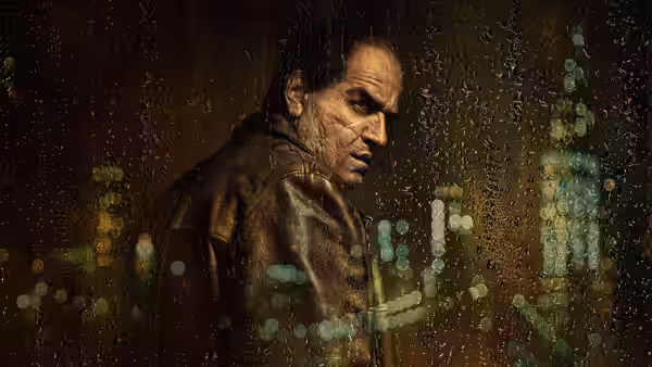 Oz Cobb in a leather jacket stands before a rain-streaked city window