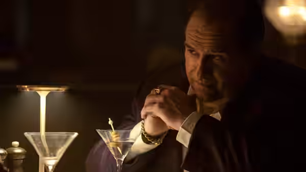 Oz Cobb sits at a bar with clasped hands beside two martini glasses
