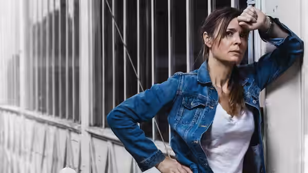 Woman in denim jacket leans against barred wall hand to forehead