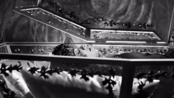 A young woman lies inside a glass-sided display surrounded by foliage
